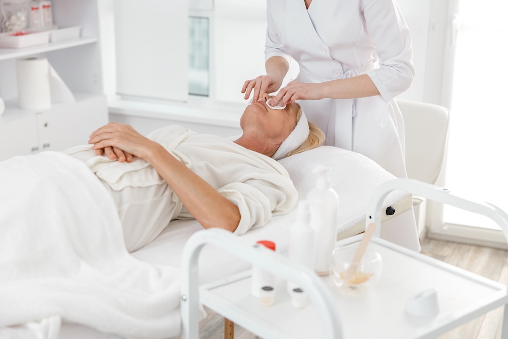 Beautiful female professional doctor is cleaning a skin of mature Caucasian blond woman in modern light cosmetology center